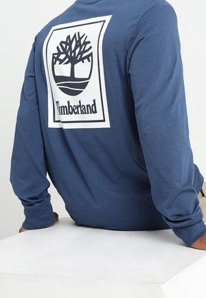 Niebieska bawełniana koszula z długim rękawem, z dużym białym nadrukiem logo drzewa i napisem "Timberland" na plecach. Gładka tekstura.