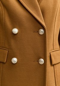 Bruine dubbelzijdige wollen jas met vier metallic decoratieve knopen en klepzakken aan elke kant.