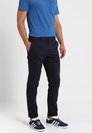 Chino blu navy con vestibilità dritta, in cotone, con tasche frontali e chiusura a bottone, abbinati a una maglietta girocollo blu e sneakers.