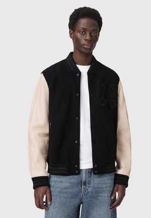 Młody mężczyzna w czarno-beżowej kurtce varsity, białej koszuli i niebieskich dżinsach, stojący na tle jednolitego jasnoszarego tła.