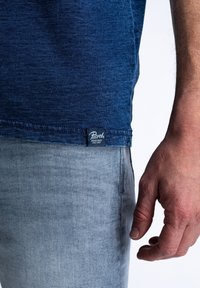 Marineblauwe katoenen T-shirt met een gestructureerd oppervlak, voorzien van een zwart Petrod-label op de zoom. Gecombineerd met lichtgrijze spijkerbroek.