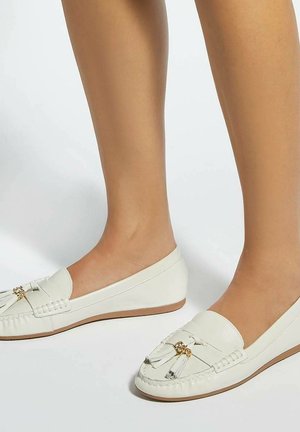 Voeten met witte leren loafers met kwastjes en gouden accenten, staand op een neutrale achtergrond.