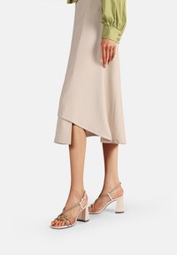 Sandali con tacco a blocco beige con una fascia frontale trasparente e dettagli metallici, abbinati a una gonna asimmetrica crema e una blusa verde.