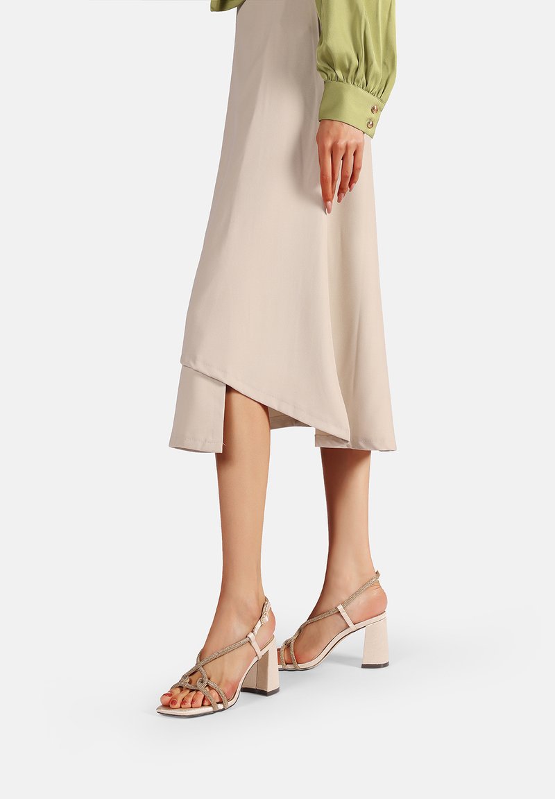 Sandali con tacco a blocco beige con una fascia frontale trasparente e dettagli metallici, abbinati a una gonna asimmetrica crema e una blusa verde.