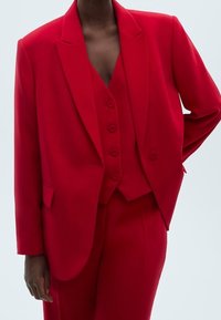 Costume rouge sur mesure avec une veste à revers crantés, un gilet ajusté à cinq boutons et un pantalon à jambes larges. Texture de tissu lisse.