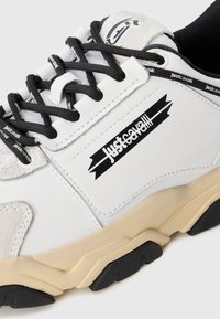 White sneaker with black laces, beige sole, and "Just Cavalli" logo on side and laces, showcasing sporty and casual design details.