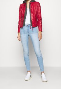 Nainen, jolla on päällään punainen nahkatakki harmaan hupparin, valkoisen T-paidan, vaaleansiniset kapeat farkut ja mustilla yksityiskohdilla varustetut valkoiset lenkkarit.