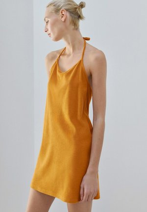 Mujer de cabello rubio recogido en un moño, vestida con un vestido naranja sin mangas y cuello halter, de pie frente a un fondo liso de color claro.