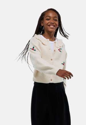 Cardigan en tricot crème avec un col à volants, une fermeture à boutons et des accents de houx brodés sur les manches. Associé à un pantalon noir.
