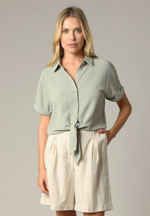 Chemise boutonnière vert clair avec manches retroussées, nouée à la taille, associée à un short crème avec des rayures verticales et des plis.