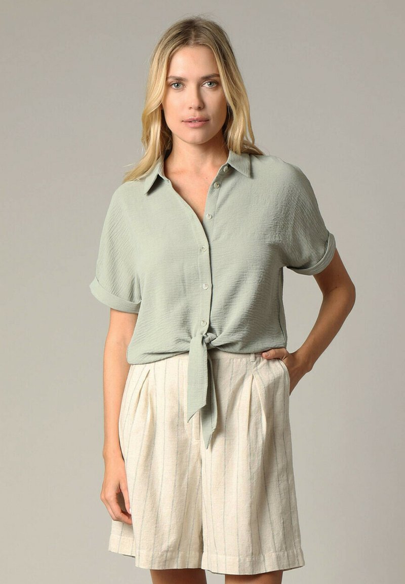 Lichtgroen overhemd met opgerolde mouwen, geknoopt in de taille, gecombineerd met crèmekleurige shorts met verticale strepen en plooien.