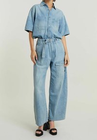 Ljusblå denimoutfit med en kortärmad skjorta med knappar och vida byxor. Har framfickor och en bälte i midjan.