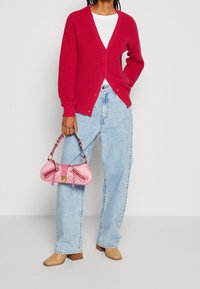Červený pletený cardigan s výstřihem do V a zapínáním na knoflíky, kombinovaný se světle modrými džínami s vysokým pasem, růžovou kabelkou a béžovými pletenými botami na blokovém podpatku.