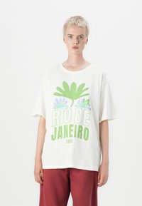 T-shirt surdimensionné blanc avec un texte graphique vert et violet "RIO DE JANEIRO 1995" agrémenté de motifs floraux. Matière en coton doux.
