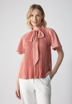 Blusa color rosa con design plissettato e un fiocco al collo. Maniche corte, texture liscia, abbinata a pantaloni bianchi.