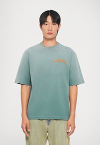 Camiseta oversize verde menta con mangas cortas que presenta un cuello redondo y un logo bordado "DSQUARED2" en naranja en el lado izquierdo del pecho.