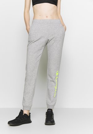 Person iført grå Champion joggingbukser med neonslogo på venstre ben, sort sports-bh og sorte Adidas sneakers, stående med hænderne i lommerne.