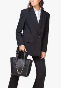 Sort struktureret håndtaske i tekstureret læder med dobbelt greb og en guld kæde accent, holdt af en person iført en sort oversized blazer og hvid skjorte.