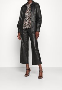 Person klädd i svart läderjacka och vida läderbyxor, blus med leopardmönster och svarta högklackade sandaler, som står mot en vit vägg.