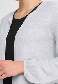 Persona con un cárdigan de manga larga gris claro sobre una blusa negra, con un tejido de punto texturizado y puños de manga ligeramente abullonados.