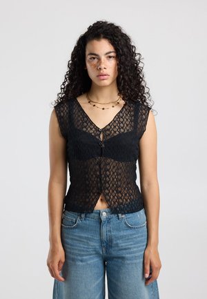 Vrouw met krullend haar en sproeten draagt een mouwloze zwarte kanten top en blauwe jeans, staand tegen een effen lichte achtergrond.