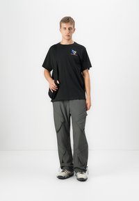 Hombre joven de pie, vistiendo una camiseta gráfica negra, pantalones cargo grises, zapatillas de color claro y llevando un bolso negro de hombro sobre fondo blanco.