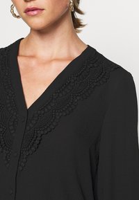Blouse noire avec des détails en dentelle délicate le long de l'encolure en V, au toucher doux et dotée d'une fermeture à boutons.