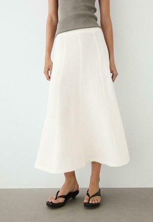 Frau trägt ein ärmelloses graues Oberteil, weißen A-Linien-Midirock und schwarze Sandaletten mit Absatz und steht vor einem schlichten Hintergrund.