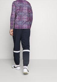 Person som bär en långärmad skjorta med lila och röda mönster, svarta byxor med vita band ovanför vristerna, och vita Nike-sneakers, står vänd bort.