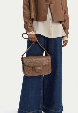 Osoba v hnědé kožené bundě, krémové blůze a širokých modrých džínách drží hnědou texturovanou koženou kabelku s horním uchem.