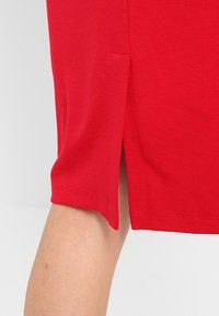 Jupe rouge longueur genou avec une fente latérale, fabriquée en tissu lisse. Elle présente un ourlet net et un design minimaliste, mettant l'accent sur la simplicité et le confort.