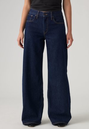 Femme portant un jean large bleu foncé et un top noir, debout, les mains relâchées le long du corps, sur un fond uni.