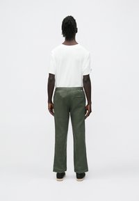 Weißes T-Shirt mit kurzen Ärmeln und olivgrüne, locker sitzende Hose mit strukturierter Oberfläche. Schwarze Clogs mit naturfarbenem Holzsohle.