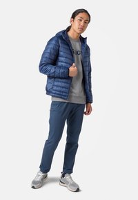 Giacca puffer blu marina con cappuccio, felpa grigia e pantaloni blu. Le scarpe sono sneaker grigie con lacci bianchi. Look complessivo casual.