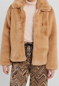 Faux-Fur-Jacke in Hellbraun mit Kragen, die eine weiche, plüschige Textur aufweist. Über gemusterten, mehrfarbigen Hosen mit Kordelzug getragen.