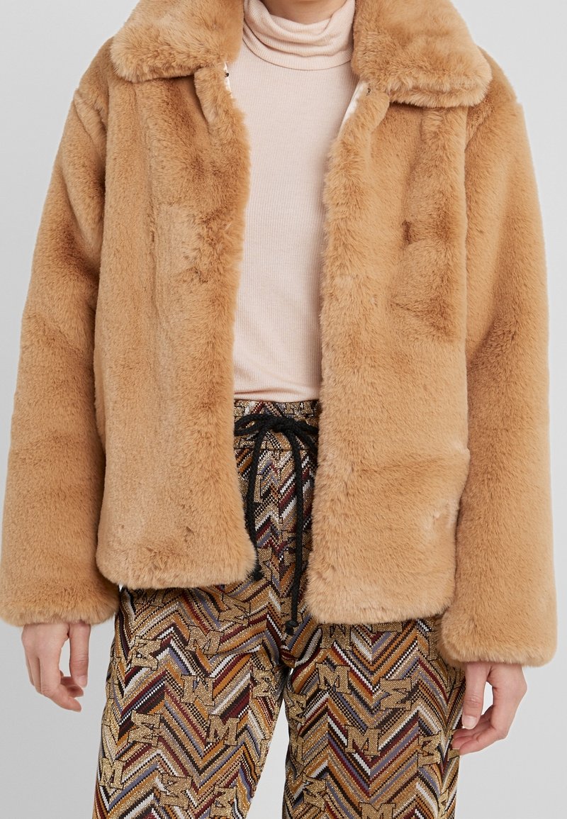 Faux-Fur-Jacke in Hellbraun mit Kragen, die eine weiche, plüschige Textur aufweist. Über gemusterten, mehrfarbigen Hosen mit Kordelzug getragen.