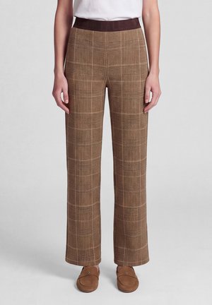 Person wearing brown plaid straight-leg pants with dark waistband, white top, and brown slip-on shoes standing on plain background.