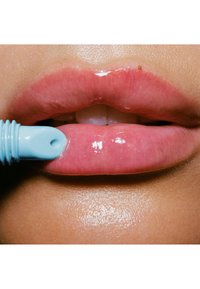Un lucidalabbra dalla formula chiara e lucida viene applicato con un applicatore azzurro. Le labbra sono piene, di un rosa chiaro e riflettente, con una consistenza liscia.