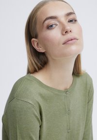 Pull en tricot vert avec un col rond, une fermeture à boutons sur le devant et des poignets côtelés. Texture douce, design ajusté et tissu légèrement texturé.