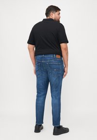 Sötétkék farmernadrág slim fit szabással, hátsó zsebekkel és barna bőr címkével a derékpánton. Fekete, strukturált póló van rajta.