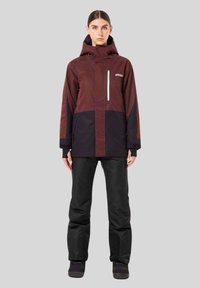 Water-resistant, two-tone jacket in maroon and black with a hood, zippered pocket, and relaxed fit, paired with black snow pants and boots.