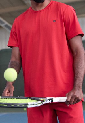 Mand iført rød skjorte og shorts, der hopper en gul tennisbold på en sort og grøn tennisketsjer indendørs.