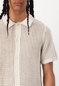 Chemise tricotée beige clair présentant un tissage ouvert respirant, des manches courtes, un col à revers et une fermeture à boutons sur le devant.