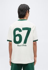 Άντρας που φοράει λευκό κοντομάνικο πουκάμισο με πράσινη επένδυση, μεγάλο πράσινο αριθμό 67 και κείμενο "Marc O'Polo" στην πλάτη.