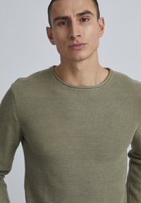 Olivgrüner, gestrickter Pullover mit Rundhalsausschnitt und gerippten Abschlüssen, der aus einem strukturierten Stoff gefertigt ist und eine taillierte Form hat.