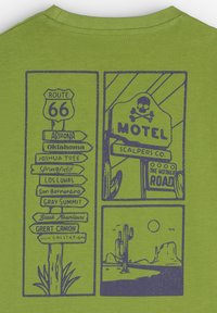 Camiseta verde con un diseño gráfico azul que presenta señales de la Ruta 66 y un logo de motel, con ilustraciones de un paisaje desértico y cactus.