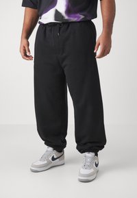 Schwarze Jogginghose mit einem verstellbaren Bund, elastischen Bündchen und einem lockeren Schnitt, kombiniert mit grau-weißen Sneakers mit navy Akzenten.