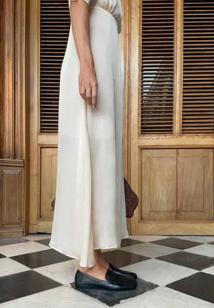 Femme portant une longue robe blanche et des mocassins noirs debout sur un sol à damier noir et blanc devant des portes en bois.