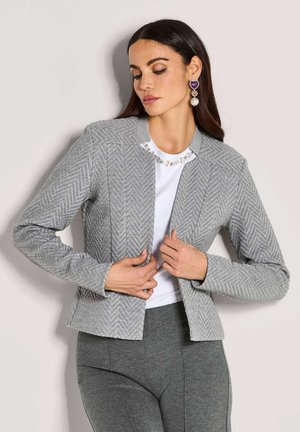 Vrouw past een getailleerde grijs visgraatblazer aan over een witte versierde top, draagt bijpassende grijze broek en opvallende oorbellen.