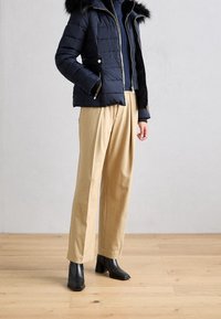 Doudoune marine avec capuche bordée de fourrure, fermeture éclair et accents dorés. Pantalon beige plissé et bottines noires à talons complètent la tenue.